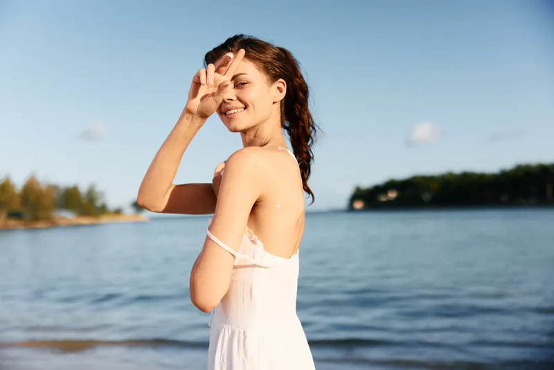 A young woman stands by a calm lake wearing a white sleeveless dress, smiling, shielding her eyes from the sun as a breeze sweeps her hair.