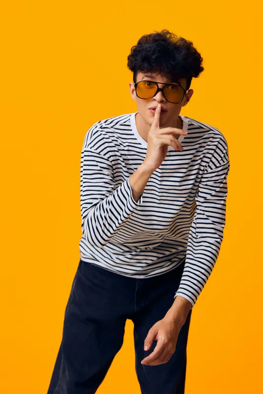 Young man in striped shirt and dark sunglasses showing silence gesture with finger on lips against vibrant orange background, casual style, modern look.