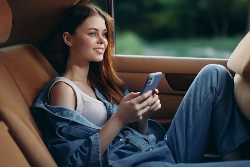 Young woman in denim outfit sitting in car, smiling while using smartphone in natural light, capturing a moment of joy and relaxation.