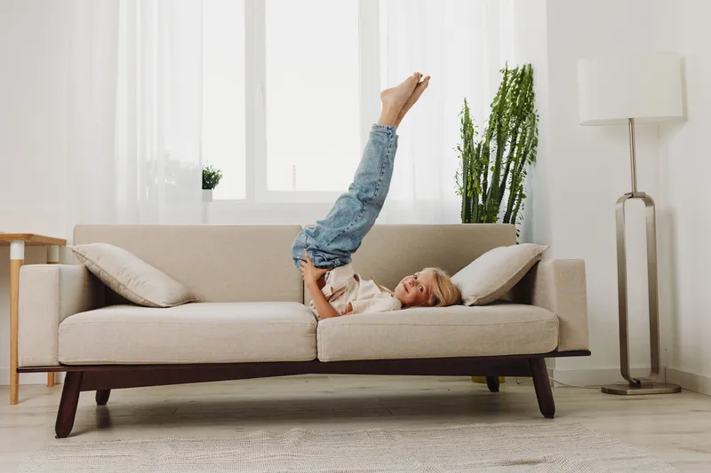 Happy child in casual clothes plays upside down on beige sofa in bright living room with plants and floor lamp. Concept of childhood joy, home comfort, and playful moments.