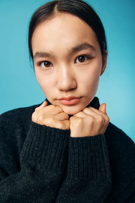 Woman with hands near face and clasped under chin in closeup portrait. Young female wearing black sweater with calm expression in studio on blue background with soft lighting.
