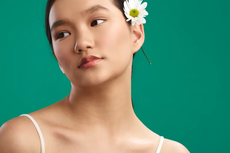 asian woman gazes softly aside in a studio portrait with a flower behind her ear on a teal backdrop, conveying calm confidence, elegance, and subtle modern style.