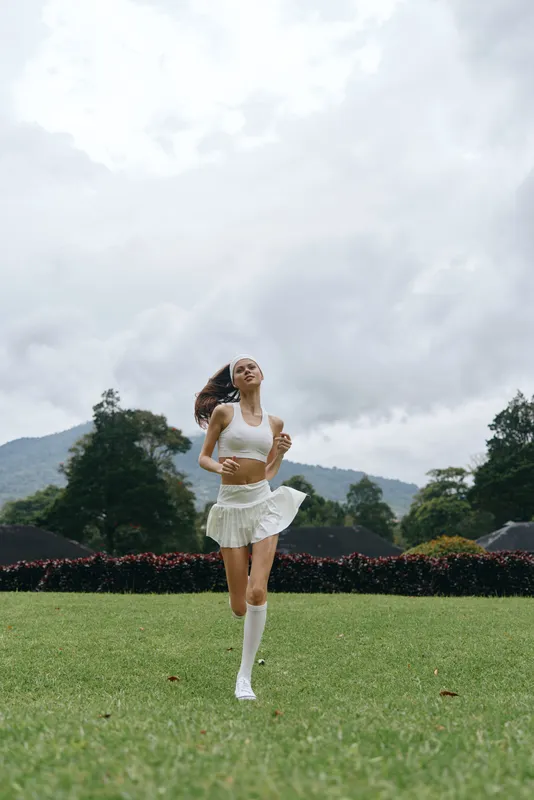 Young athletic woman in white sportswear joyfully running on a grassy field with a cloudy sky, embodying fitness and vitality in a natural setting