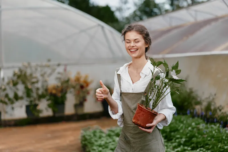 Cheerful young woman in overalls showcasing a potted plant in a greenhouse, emphasizing sustainability and a love for gardening