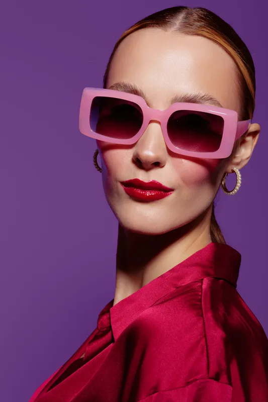 Stylish woman with pink sunglasses and red lipstick poses confidently against purple background. Fashion portrait with bold makeup and trendy accessories for modern style concept.