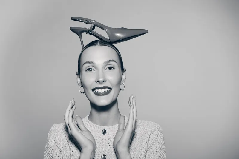 Smiling caucasian woman with pale skin holding shoe on head wearing knitted sweater and hoop earrings in studio. Concept of fashion and humor.