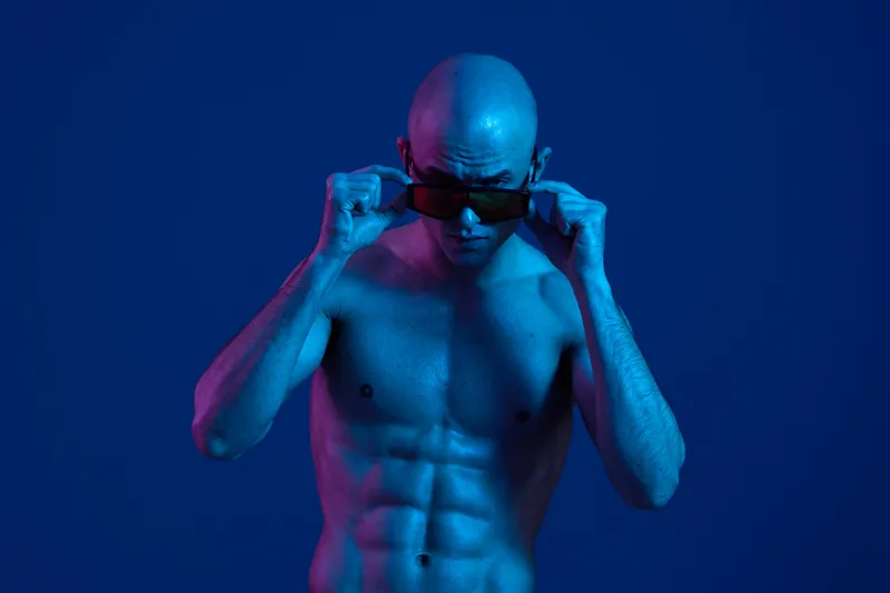 Muscular man posing confidently in sunglasses, illuminated by vibrant blue and purple lighting, showcasing strength and style in a unique way.