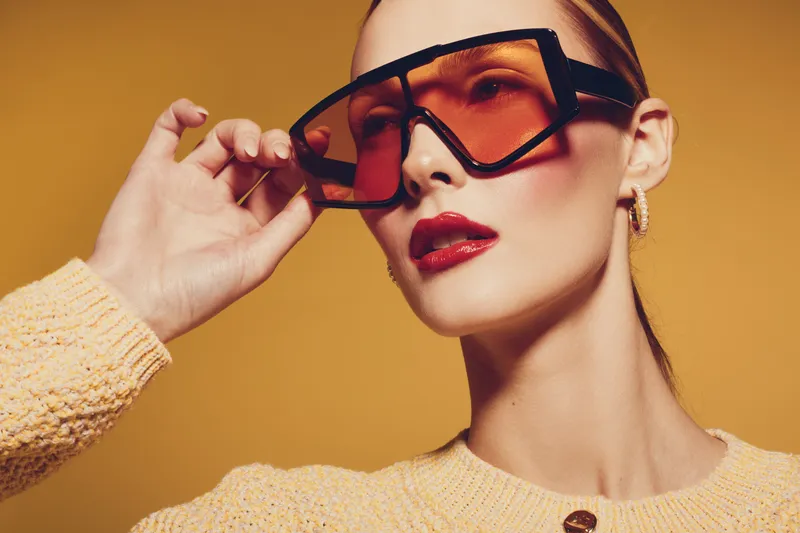 Stylish woman with slicked hair wearing oversized tinted sunglasses and red lipstick poses against yellow background. Fashion concept with modern accessories and confident expression.