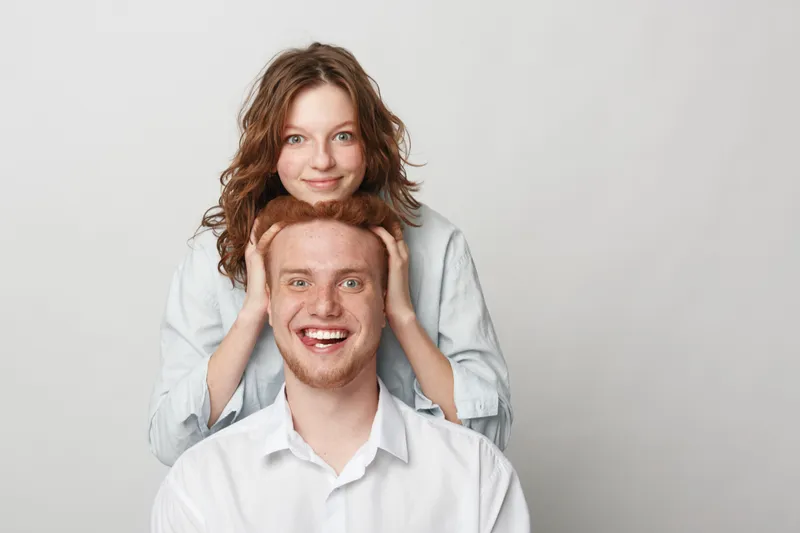 Smiling young woman with curly hair stands behind happy redhead man in white shirt. Studio portrait on grey background. Concept of friendship, joy, and positive emotions.