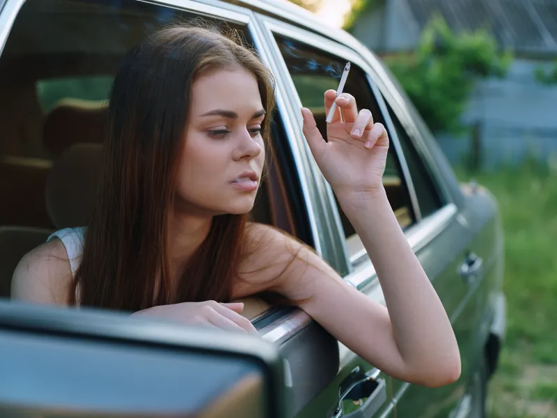 Thoughtful young woman smoking in a car, looking out the window, with a contemplative expression against a natural background.