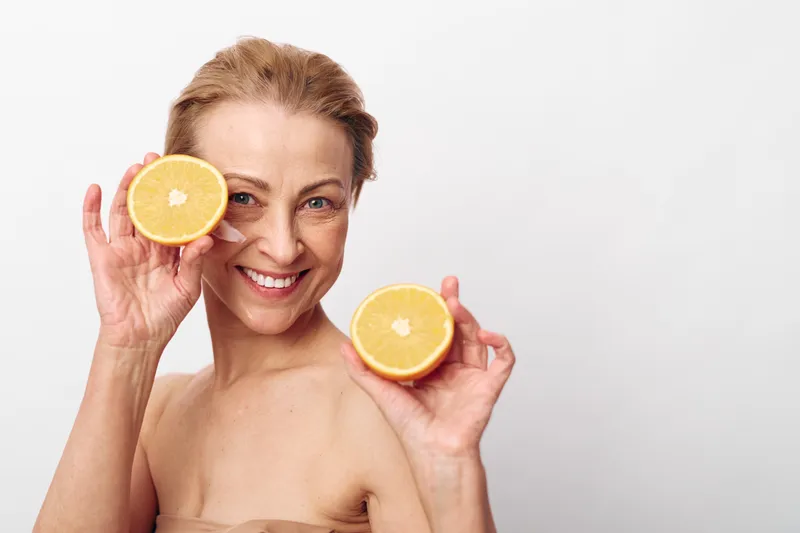 Smiling middle aged woman holds two halves of fresh orange fruit near her face, showing healthy skin and natural beauty on white background. Concept of wellness, skincare, and vitamin health.
