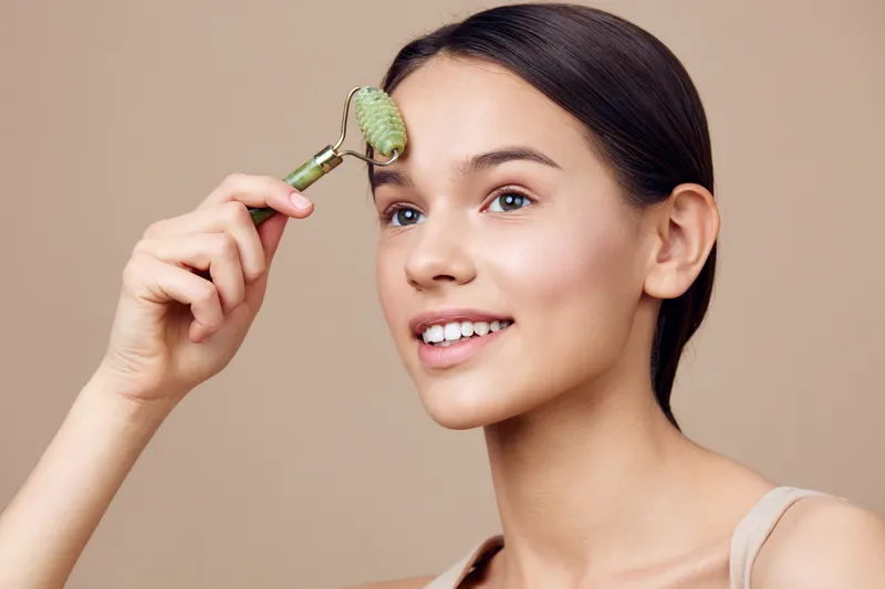 Young woman using a jade roller on her face for skincare, showcasing a natural beauty routine with a relaxed expression in a neutral background