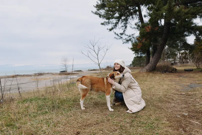 Woman in a winter coat kneels beside a dog on a leash by the coast, embracing the moment in a quiet park with bare trees and calm water nearby.
