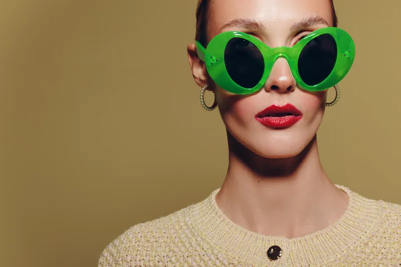 Stylish woman wearing large green sunglasses and red lipstick poses against beige background. Fashion portrait with trendy accessories and bold makeup concept.