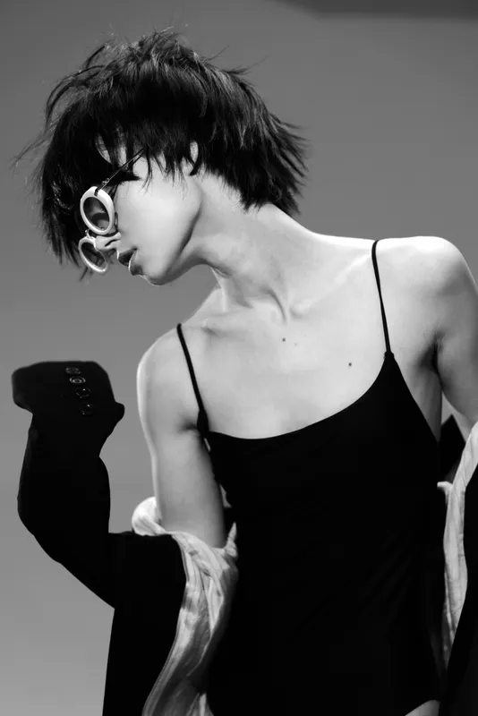 Black and white portrait of a stylish woman with short hair wearing glasses and a black dress showing elegant posture and attitude.