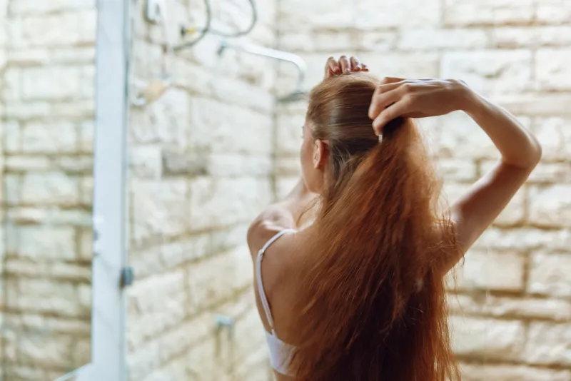 woman with long hair in white top brushing hair in modern bathroom with light stone wall and glass shower door, natural light, morning routine, self-care