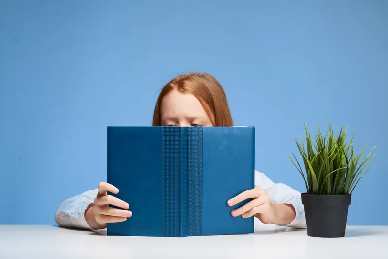Рыжеволосая девушка сидит за столом с книгой в руках и учит уроки