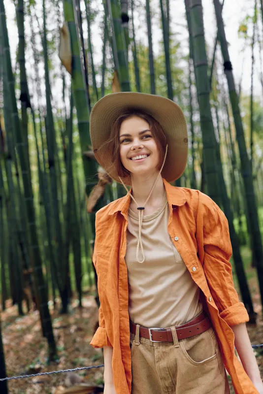 Woman smile hat bamboo forest outdoors portrait travel: young woman in orange shirt and widebrim hat smiling among tall bamboo stalks, casual travel lifestyle, nature escape and relaxed mood.