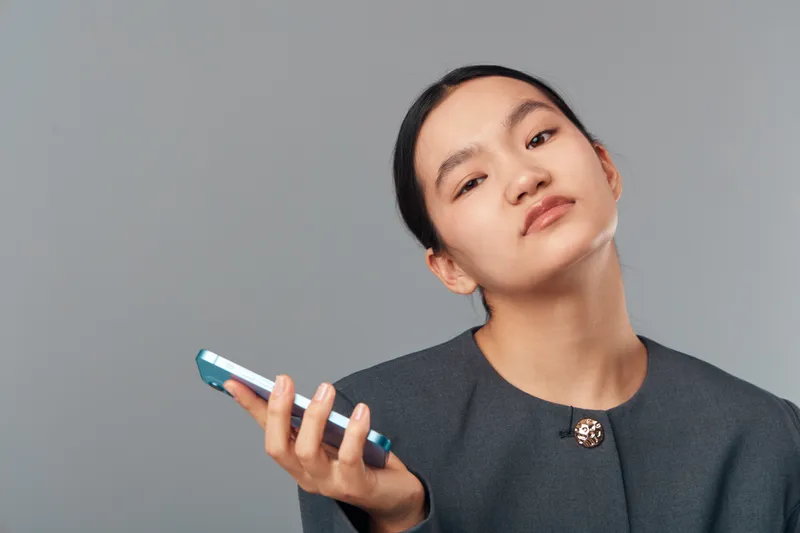 Young woman holding phone with serious expression, posing in studio with minimalist grey background and wearing dark blazer. Portrait of thoughtful female with modern device in hand.