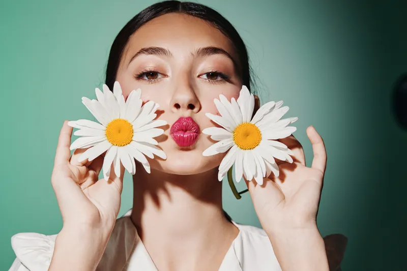 Young woman in a minimalist outfit holds two white daisies near her face while puckering her lips. Studio shot with pastel green background forms a beauty and creative fashion concept.