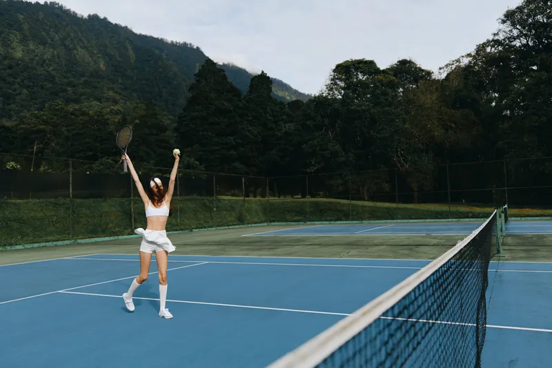 Joyful young woman celebrating a tennis victory on a sunny day, wearing a sporty white outfit with a backdrop of mountains and green trees