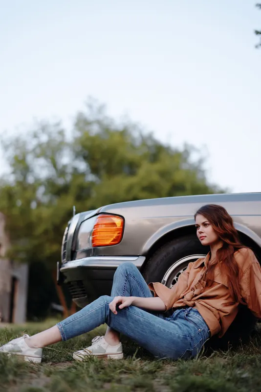 Young woman sitting casually beside a vintage car, wearing a brown shirt and jeans, exuding a relaxed and stylish vibe, set in a natural outdoor background.