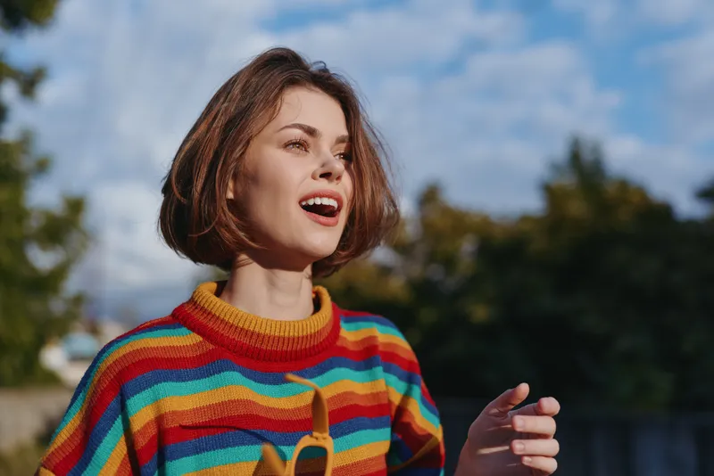 Woman smiling in a colorful striped sweater with short hair bob, outdoors under a blue sky. Joyful young adult holding sunglasses, casual outfit and lifestyle portrait for travel and fashion.