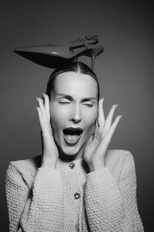 Young white woman with light skin closes eyes and screams holding hands to face with shoe on her head in black and white photo.