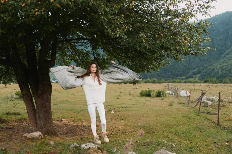 Young woman in white clothes stands under tree in countryside holding gray blanket with arms raised, enjoying fresh air and natural landscape with mountains in background.