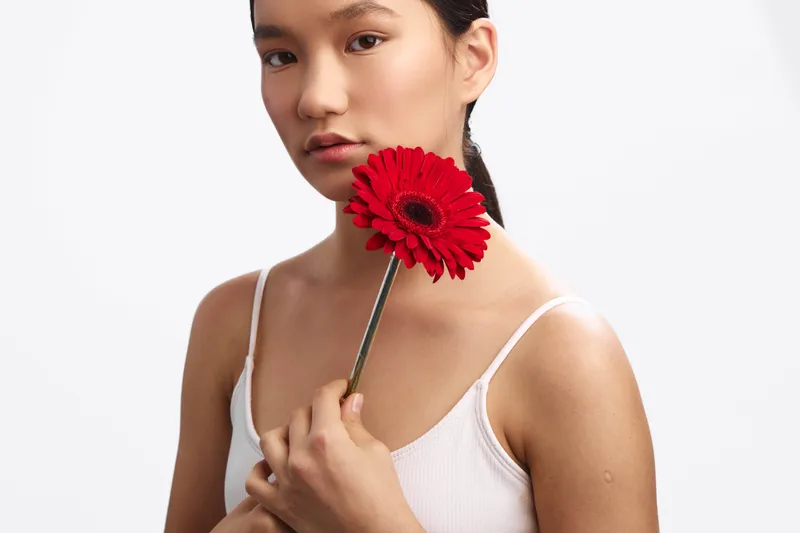 Asian woman stands gracefully against a plain backdrop, holding a red flower near her chest. She looks softly at the camera with a calm, thoughtful expression, conveying subtle elegance and a serene,