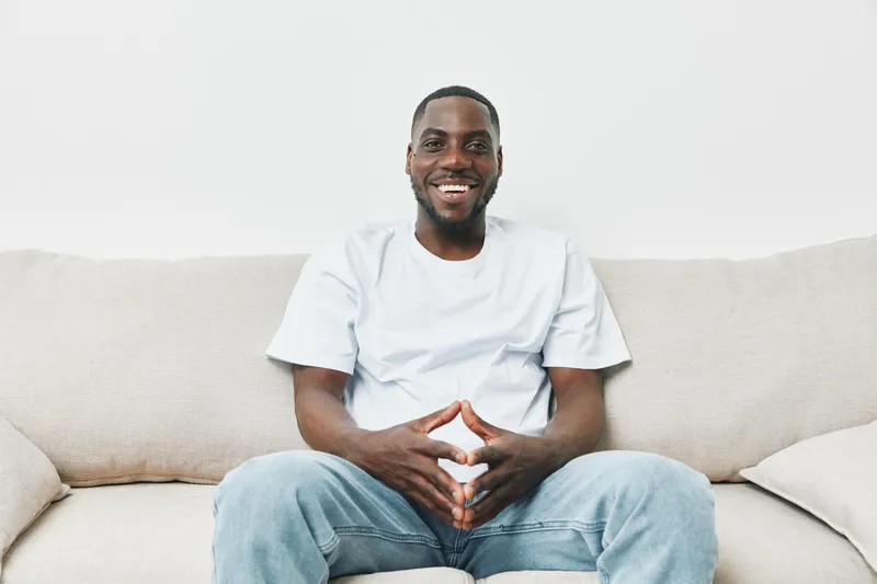 Happy African American man sitting on a couch, wearing casual white t shirt and jeans, expressing joy and comfort in a cozy home setting