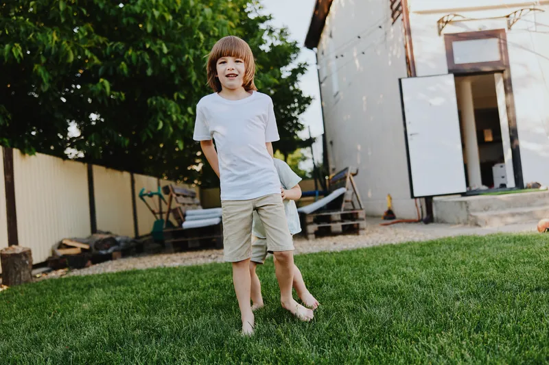 Happy boy playing outdoors in the garden, enjoying summer vibes and freedom. He wears a casual white t-shirt and shorts, standing barefoot on the grass.