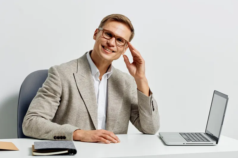 Smiling man in business casual attire sitting at a desk with a laptop, looking confident and professional, isolated on white background, suitable for corporate, technology, or office concepts.