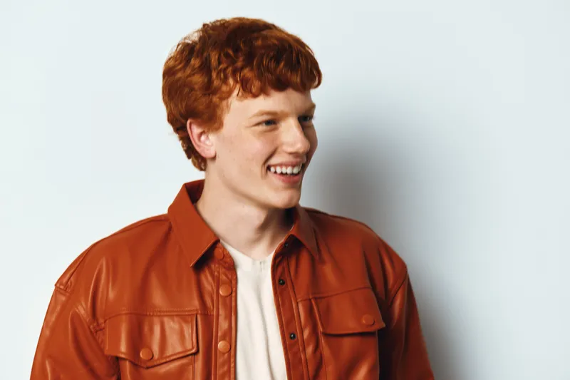 Young man with red hair wearing brown leather jacket smiling and looking aside on white background. Casual style portrait with happy expression and natural lighting.