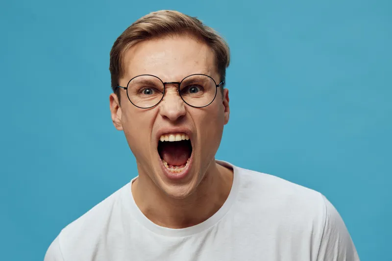 frustrated angry young caucasian man with glasses shout yell with open mouth on blue background white tshirt mental health and gadgets concept