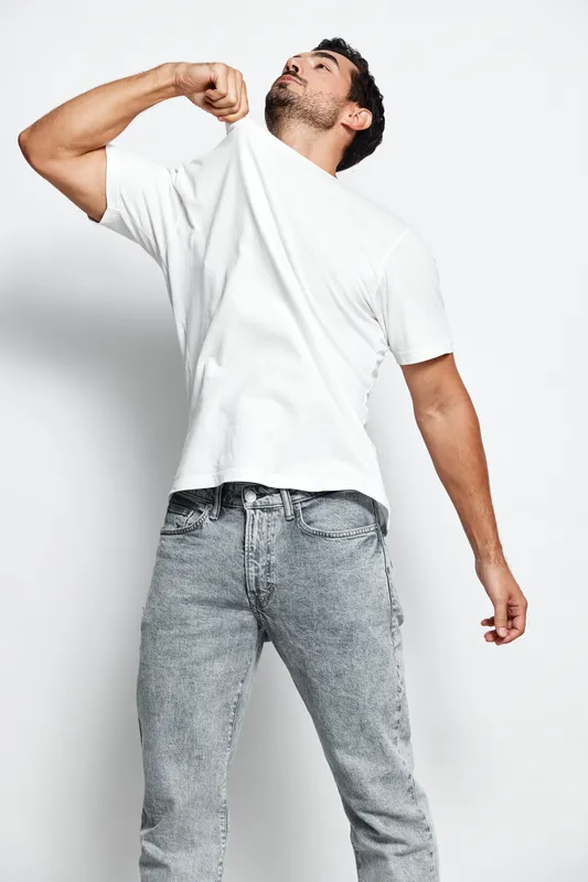 Handsome man pulling up his white shirt, showcasing a confident and relaxed demeanor against a neutral background, capturing a moment of self-expression in style.