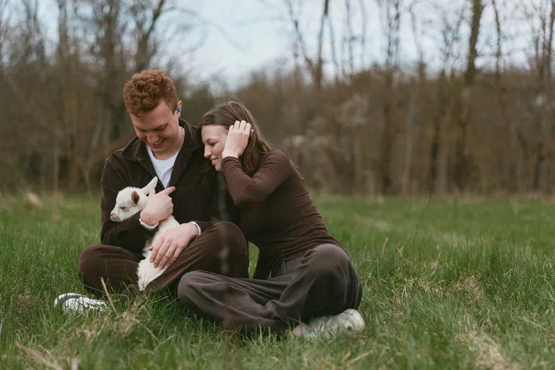 Loving couple outdoors with a goat kid in a grassy field, moment of companionship and gentle play. Casual clothing, soft natural light, serene mood, rural setting, heartwarming interaction.