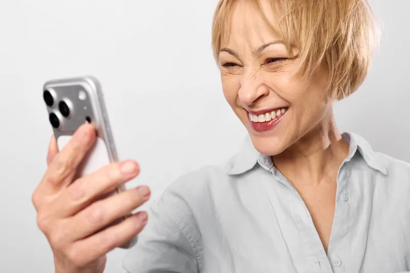 Elderly woman smiles while holding smartphone, laughing with eyes closed in a bright indoor studio, concept of friendly communication and everyday technology