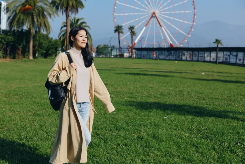 Woman park coat backpack walking outdoors in sunny weather beside a ferris wheel, urban leisure scene with greenery, casual fashion and photography vibe in a daylight setting