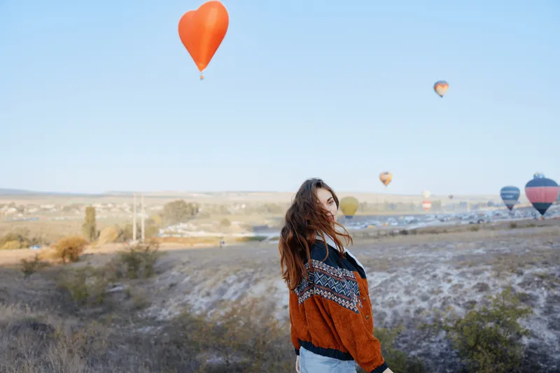 Женщина стоит на живописном поле, а вдалеке под ясным голубым небом виден воздушный шар в форме сердца