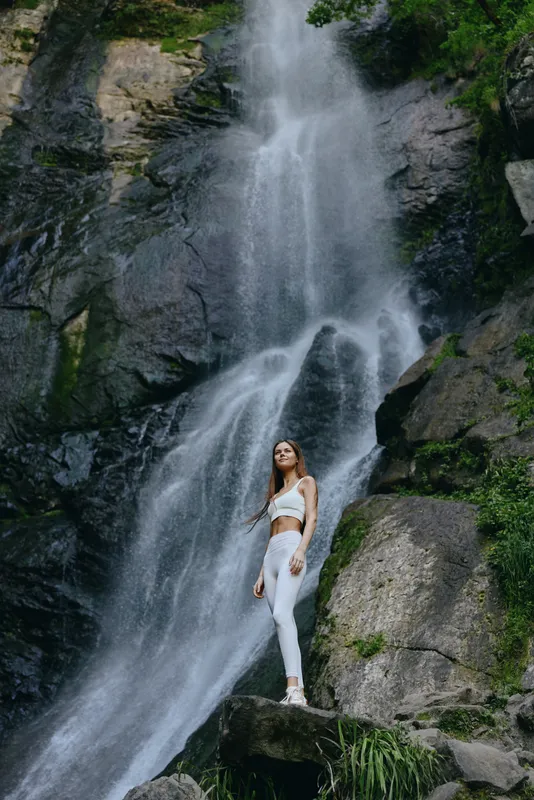 woman standing near waterfall wearing white activewear, outdoors, rocky landscape, serene nature, healthy lifestyle, adventure and fitness.