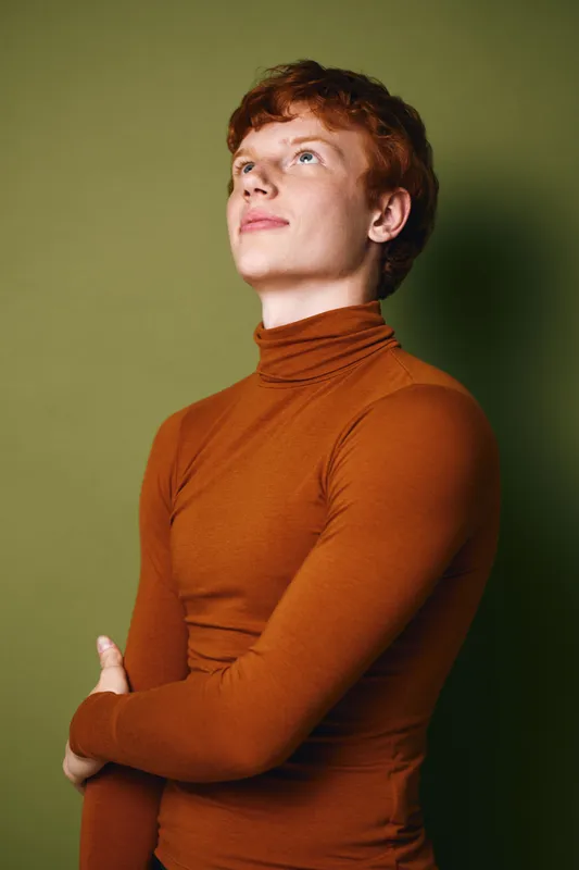 Young man with red hair wearing orange turtleneck sweater stands with arms crossed looking up against green background. Casual style portrait of confident male model with thoughtful expression.