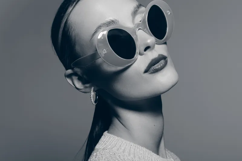 Portrait of a young white woman with pale skin wearing large round sunglasses and hoop earrings. Studio fashion shot with monochrome lighting and confident expression.