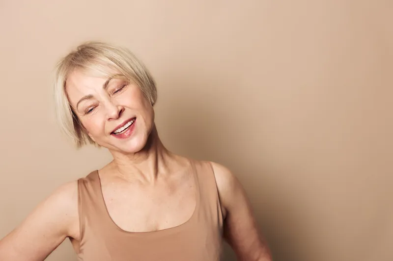 Smiling mature woman with short blonde hair wearing beige tank top poses with closed eyes against soft beige background. Concept of happiness, natural beauty, and relaxed mood in studio.