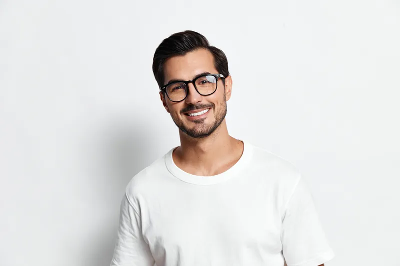 Smiling young man wearing glasses and a white t-shirt looks at the camera with a cheerful expression against a plain solid background. People lifestyle concept