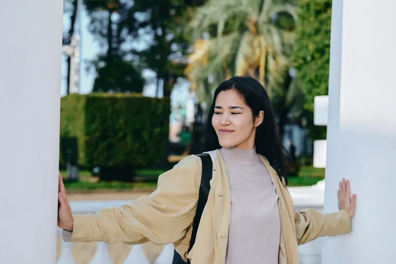 Woman stands between white pillars outdoors, wearing a beige jacket and light sweater, eyes relaxed, serene pose, soft sunlight, peaceful urban park atmosphere during a casual stroll