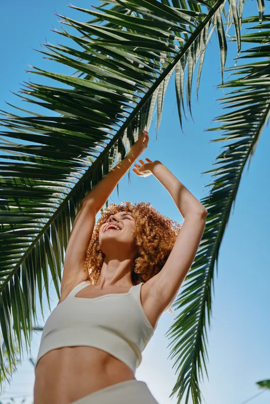 Smiling woman with curly hair stretches joyfully under green palm leaves on a bright sunny day with clear blue sky background, radiating happiness and freedom.