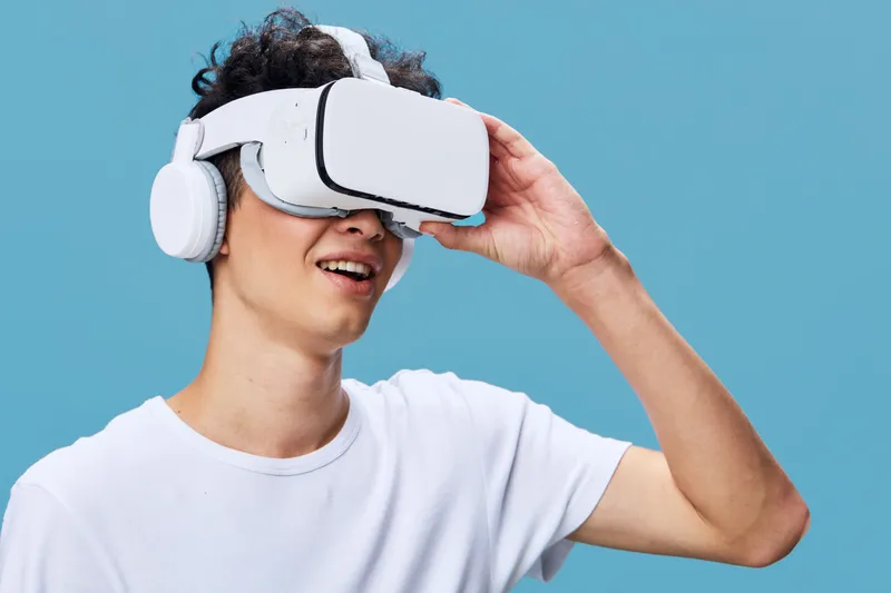 Young man wearing virtual reality headset and white headphones exploring VR technology against a solid blue background, enjoying immersive digital experience.