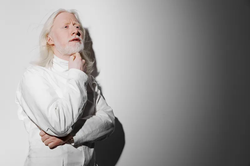 Портрет демонстрирует пожилого мужчину с поразительными седыми волосами и бородой, одетого в безупречный белый костюм