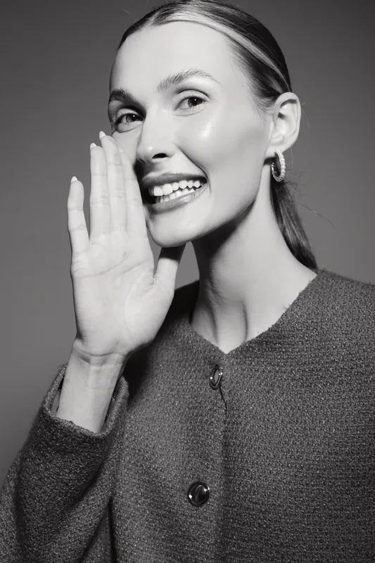 Smiling Caucasian woman with fair skin and pulled back hair whispers with hand near mouth. Studio portrait with soft lighting and casual clothing. Concept for communication and secrets.
